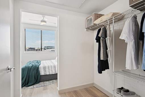 A white bedroom with a bed, a window with blinds, a clothing rack with clothes, and a pair of shoes on the floor.