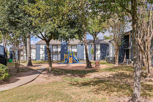 A playground with a slide is surrounded by trees and a building in the background.