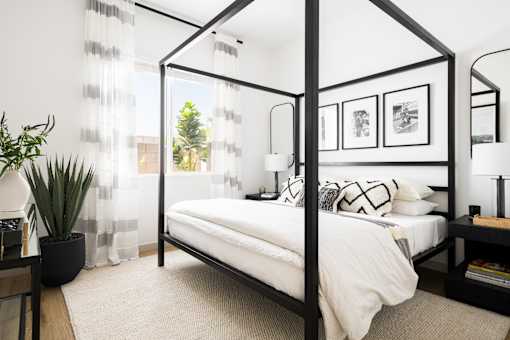 A white bed with a black frame is in a room with a white wall and a window with white curtains.