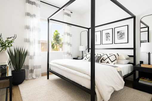 A white bed with a black frame is in a room with a white wall and a window with white curtains.