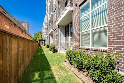 A long, narrow, green lawn separates a brick building from a wooden fence.