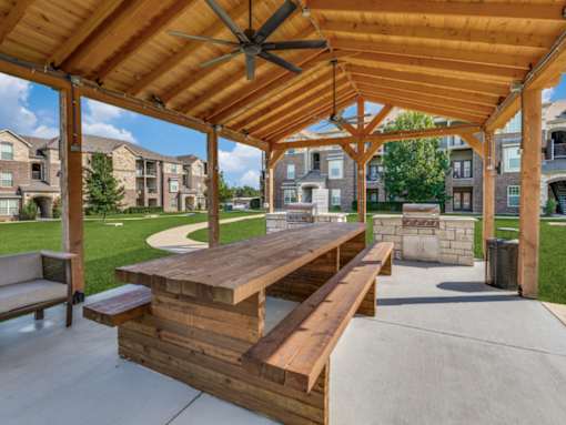 A wooden pavilion with a bench and a table in the foreground and apartment buildings in the background.