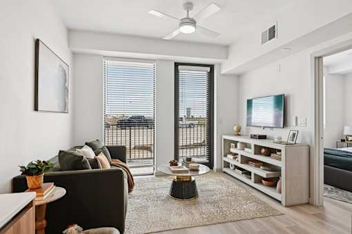 A living room with a brown couch, a coffee table, a television, and a balcony with a view of the city.