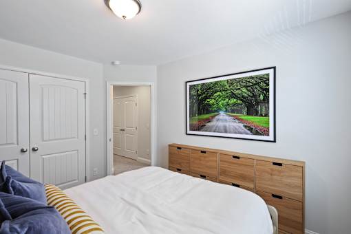 A bedroom with a white bed and a painting of a tree-lined road on the wall.