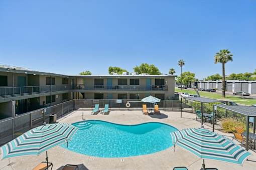 A large swimming pool with lounge chairs and umbrellas is surrounded by apartment buildings.