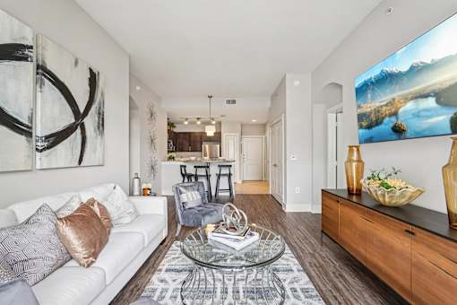 A modern living room with a white sofa, a glass coffee table, and a large painting on the wall.