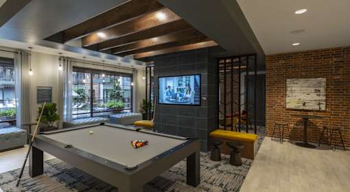 A pool table is in the middle of a room with a brick wall and a television.
