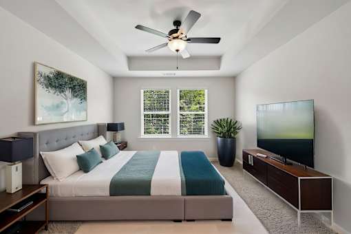 A bedroom with a bed, a ceiling fan, a window with blinds, a painting, a potted plant, a TV, and a wooden cabinet.