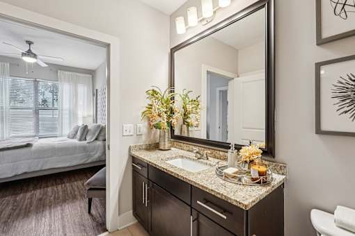 A bathroom with a large mirror, a sink, and a bedroom with a bed and a window.