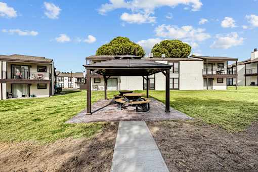 A gazebo sits in the middle of a grassy area with apartment buildings in the background.