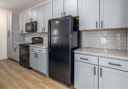 A black refrigerator stands in a kitchen with white cabinets and a granite countertop.
