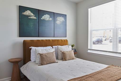 A bedroom with a bed, pillows, and a painting of clouds on the wall.