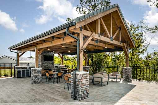 A wooden pavilion with a stone pillar and a fireplace is surrounded by chairs and tables.