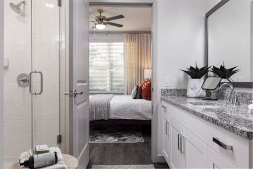 A bathroom with a white shower, a bed with a white blanket, a fan, and a mirror.