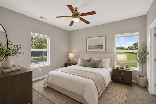 A bedroom with a large bed, a ceiling fan, and a view of a green landscape through the windows.