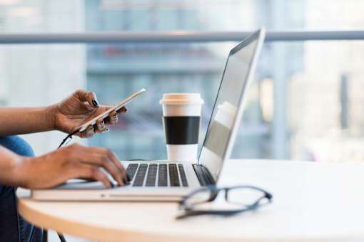 A person is using a laptop and holding a smartphone while sitting at a table with a cup of coffee.