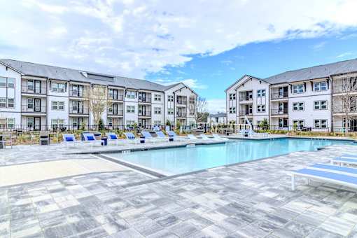 A large swimming pool surrounded by a tiled floor and lounge chairs in front of apartment buildings.