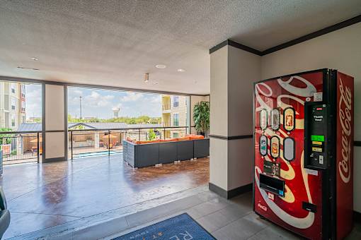 A Coca-Cola vending machine is in a room with a view of a balcony and buildings outside.