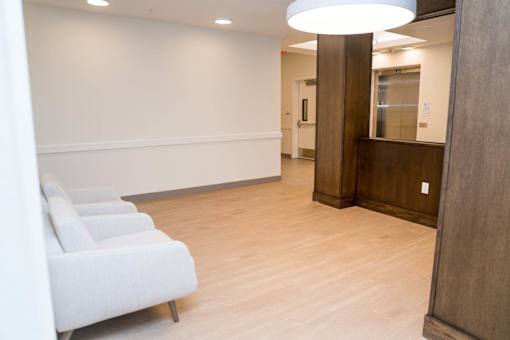 A hallway with a row of white chairs on the left and a wooden door on the right.