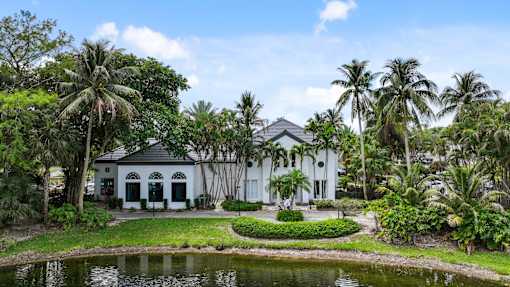 A white house with a black roof is surrounded by palm trees and a pond.