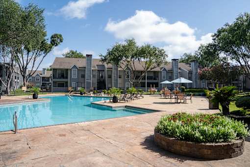 A pool surrounded by a patio and trees with a building in the background.