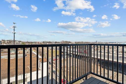 A balcony with a black railing overlooks a parking lot and a road.
