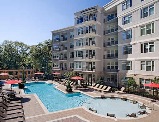 A large swimming pool surrounded by lounge chairs and umbrellas in front of a multi-story apartment building.
