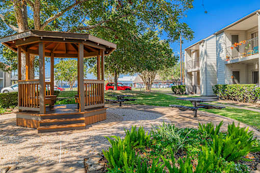 A gazebo sits in the middle of a grassy area with a bench and a tree nearby.