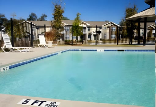 a swimming pool with two white chairs and a fence with apartments in the background