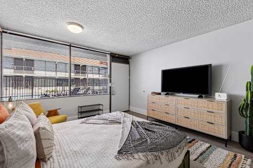 A bedroom with a bed, a couch, a television, and a window with a view of a building.