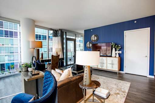 A blue chair sits in a living room with a white couch and a table with a lamp on it.