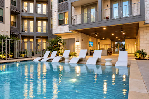 A swimming pool in front of a building with white chairs around it.