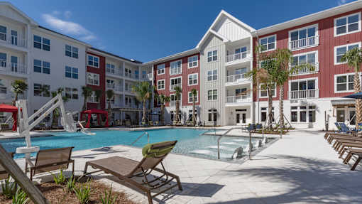A pool area with lounge chairs and a fountain in front of apartment buildings.