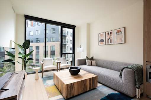 A modern living room with a grey couch, a wooden coffee table, and a large window overlooking a cityscape.