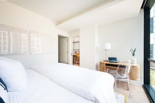 A white bed with a white pillow is in a room with a desk and chair.