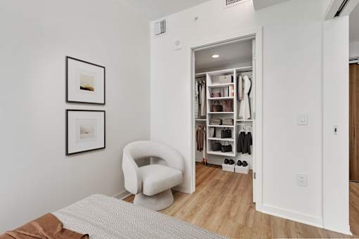 A white chair sits in a room with a white wall and a closet with clothes.