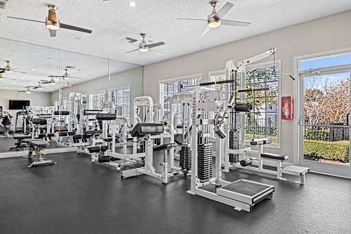 A spacious gym with a variety of equipment and a view of a fence and trees outside the window.