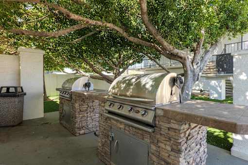 A large outdoor grill is built into a stone wall under a tree.