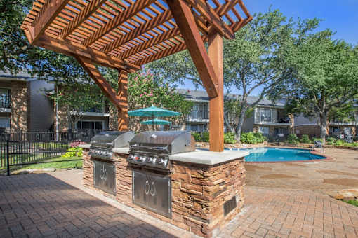 A wooden pergola over an outdoor kitchen with a grill and a pool in the background.