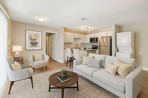 A living room with a grey couch, two chairs, a coffee table, and a kitchen area in the background.