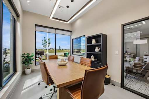 A conference room with a long table and chairs, a potted plant, and a view of the outdoors through large windows.