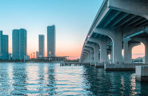 A city skyline is reflected in the water under a bridge.