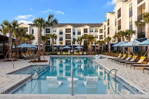 A swimming pool surrounded by lounge chairs and umbrellas in front of a building with palm trees.