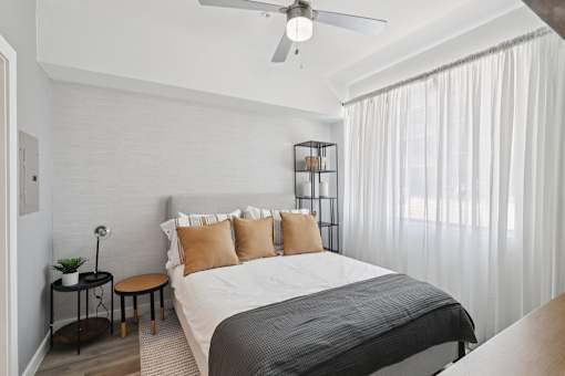 A bedroom with a bed, pillows, a ceiling fan, and a small table with a plant on it.