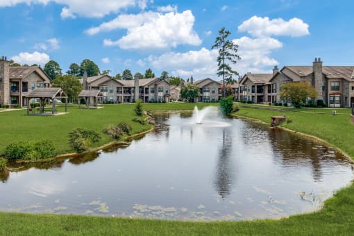 a pond with a fountain in the middle of a green field with houses