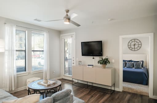 A living room with a grey couch, a round table, a television, and a bed with a blue comforter.