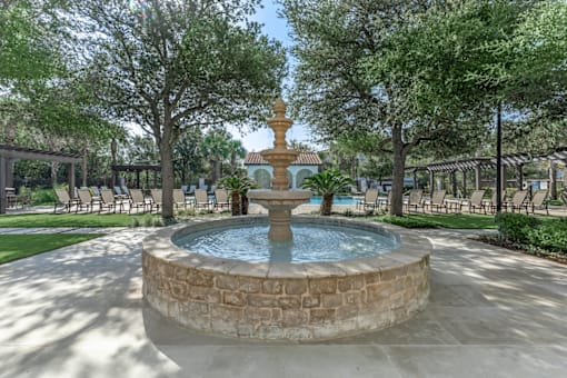 A fountain in the middle of a park with trees and chairs around it.