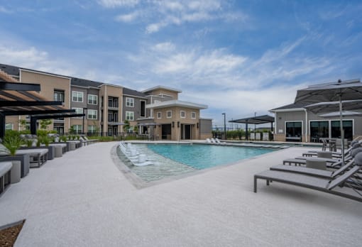 A swimming pool surrounded by sun loungers and umbrellas in front of apartment buildings.