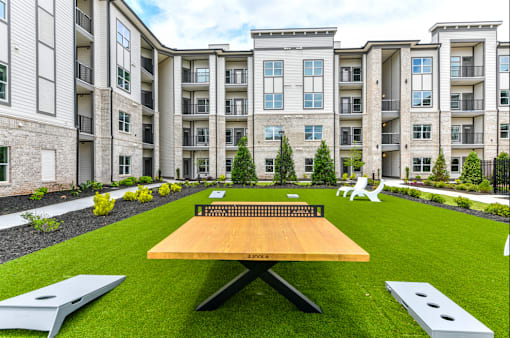 A wooden picnic table sits in the middle of a grassy area in front of apartment buildings.