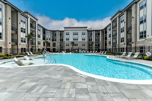 A large swimming pool in front of a building with lounge chairs around it.